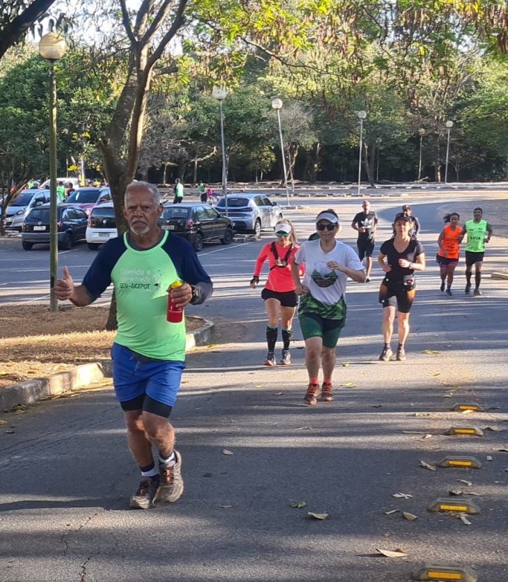 A imagem mostra um grupo de pessoas praticando corrida em uma área arborizada e sombreada do Campus Pampulha da UFMG. Algumas pessoas estão correndo na pista enquanto outras caminham, com árvores e postes de iluminação ao fundo. #ParaTodosVerem Foto: Carlos Alberto/criador do grupo “Femegê”