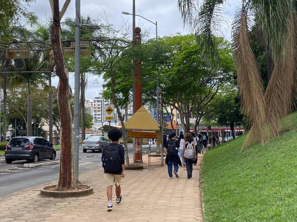 A foto é da entrada da Av. Antônio Carlos do Campus Pampulha da UFMG. Nela, há diversos alunos de costas caminhando pela calçada. Há alguns carros passando e à direita um espaço com grama.