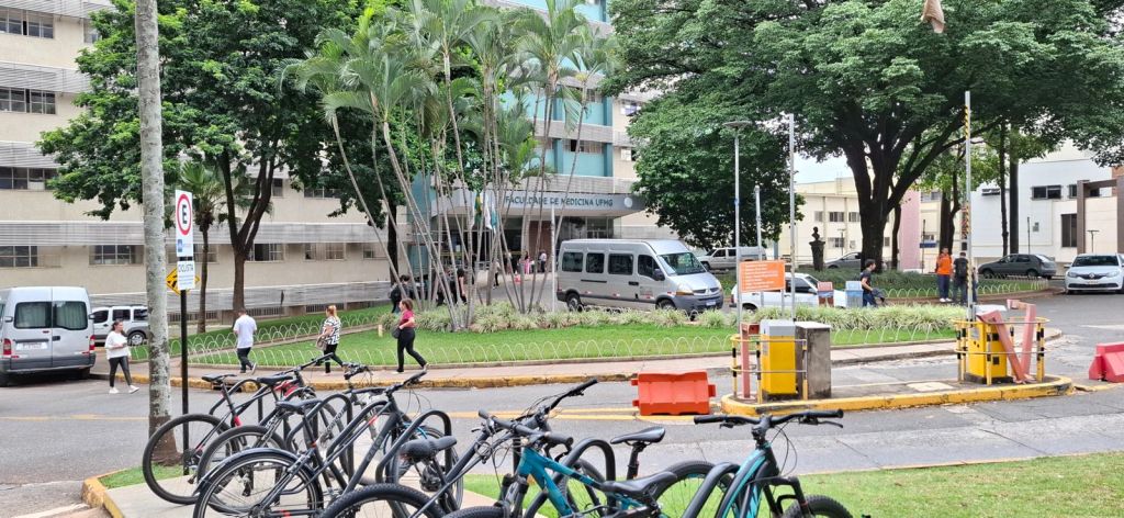 Foto da portaria do Campus Saúde da UFMG. Nela, tem um bicicletário com muitas bicicletas estacionadas e algumas pessoas caminhando. Tem uma van estacionada ao centro e algumas árvores.