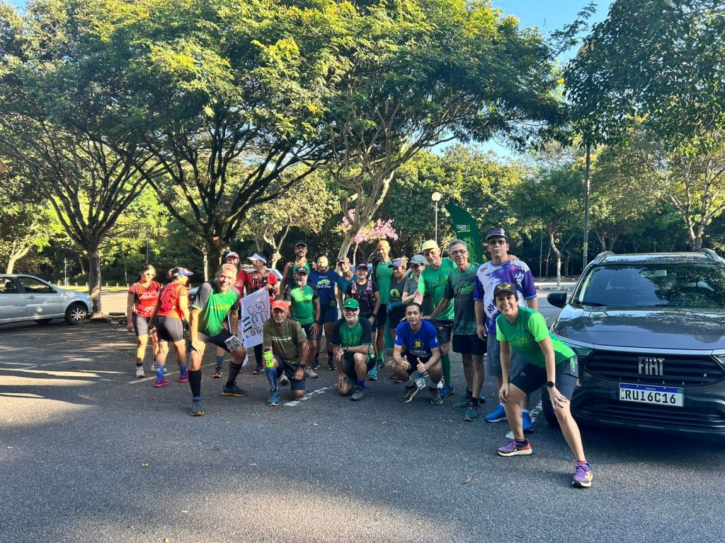 A imagem mostra um grupo de 20 pessoas reunidas em um estacionamento arborizado, em um dia ensolarado. A maioria está usando camisetas de cores vibrantes, como verde, vermelho e azul, e calções ou leggings. Algumas pessoas estão agachadas na frente, enquanto outras estão em pé, sorrindo para a câmera. Duas pessoas seguram uma bandeira com o escrito "Fêmege". À direita do grupo, um carro Fiat cinza escuro está estacionado, e há árvores verdes ao fundo. 

#ParaTodosVerem

Foto: Carlos Alberto/criador do grupo “Femegê”