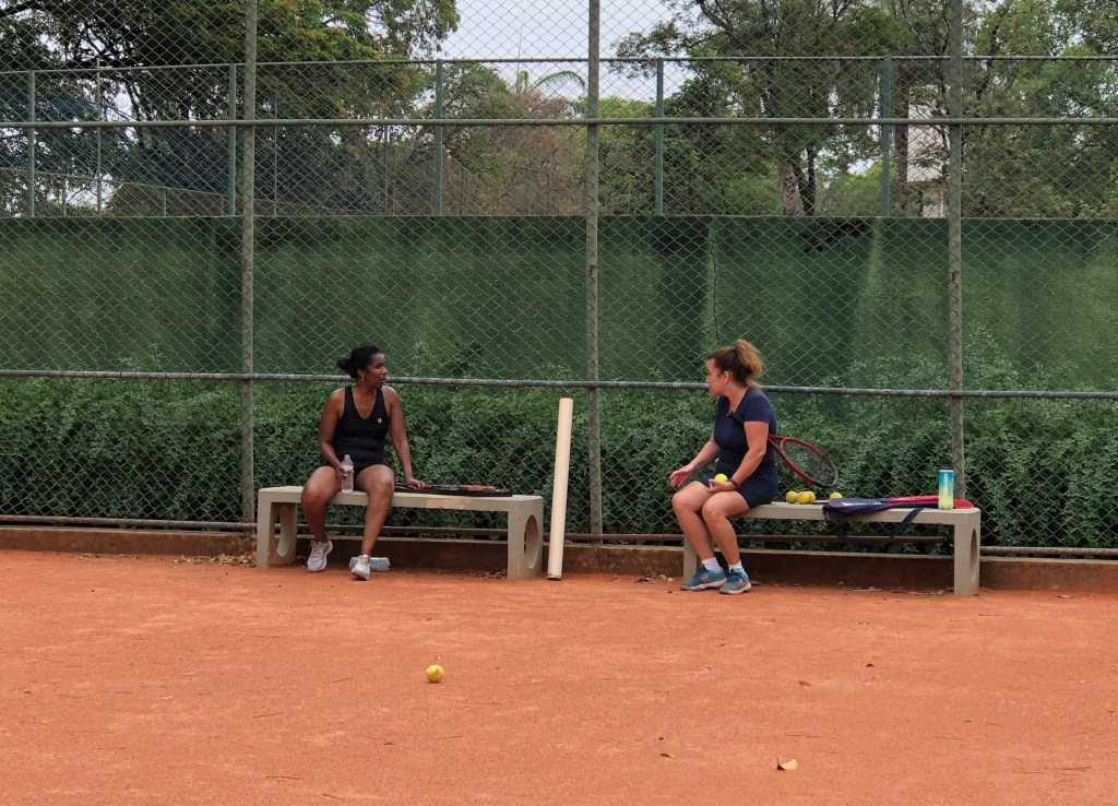 #ParaTodosVerem: Duas mulheres conversando sentadas em bancos de uma quadra de tênis.