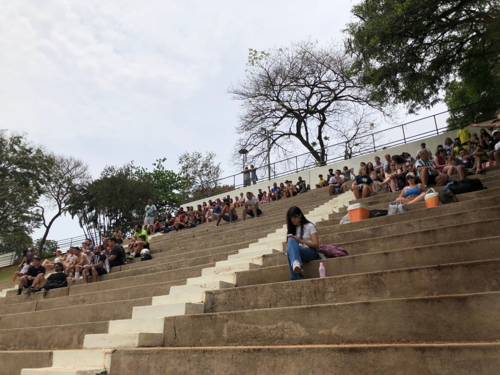 #ParaTodosVerem: Um aglomerado de pessoas sentado em uma arquibancada a céu aberto para assistir a um jogo de futsal.