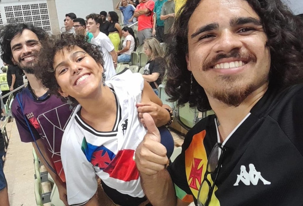 Três pessoas com a camisa do Vasco estão dentro da arquibancada de um estádio de futebol. Da esquerda para a direita: Um homem branco, Samuel, de cabelo preto médio, com idade entre 20 e 25 anos, vestido com uma camisa roxa do Vasco da Gama, ao seu lado, uma mulher parda, com cabelo curto, idade entre 20 e 23 anos, com uma camisa branca com listra diagonal em cor de arco-íris do Vasco e, por último, um homem pardo, de com cabelos próximos aos ombros, com idade entre 20 e 23 anos, vestido com uma camisa preta e listra diagonal de arco-íris do Vasco da Gama.