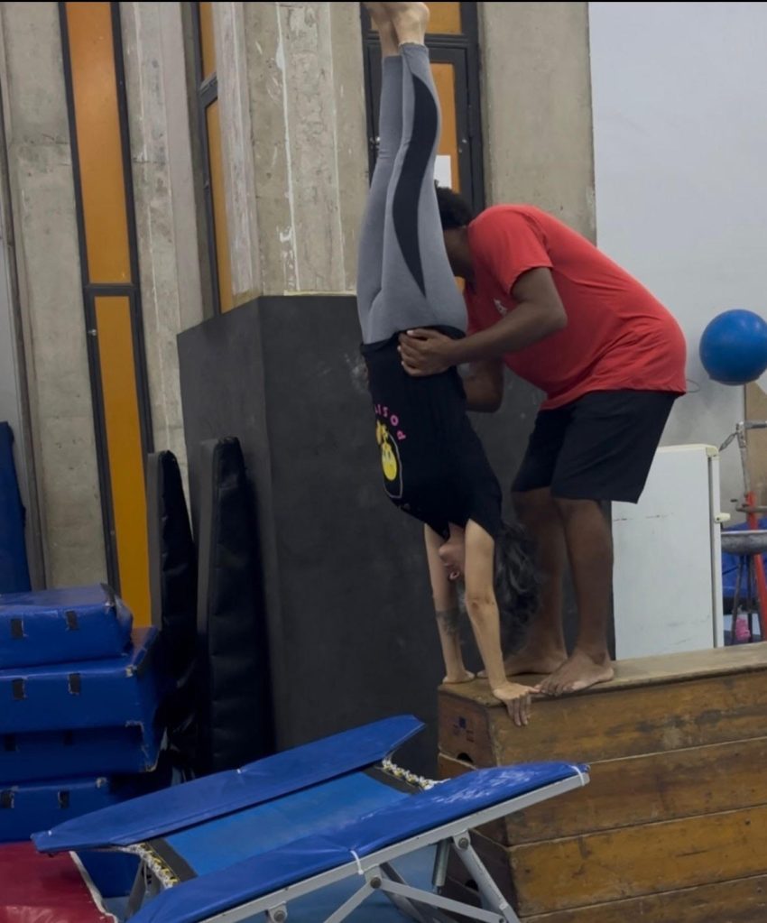 Mulher com camisa preta e calça legging cinza realiza uma postura de cabeça para baixo, apoiada nas mãos, enquanto é auxiliada por um homem, que usa uma camisa vermelha e bermuda preta. Eles estão sobre um tablado de madeira, com um trampolim azul à frente