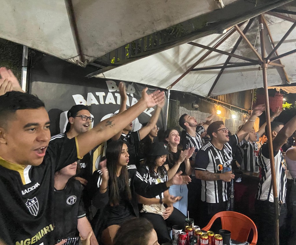 12 pessoas estão torcendo para o Atlético Mineiro em meio a um bar. 10 delas estão de pé e 3 estão sentadas. Todos estão vestidos de Galo, cantando e gesticulando. Ao centro da foto, está escrito Bar Batata Raiz.