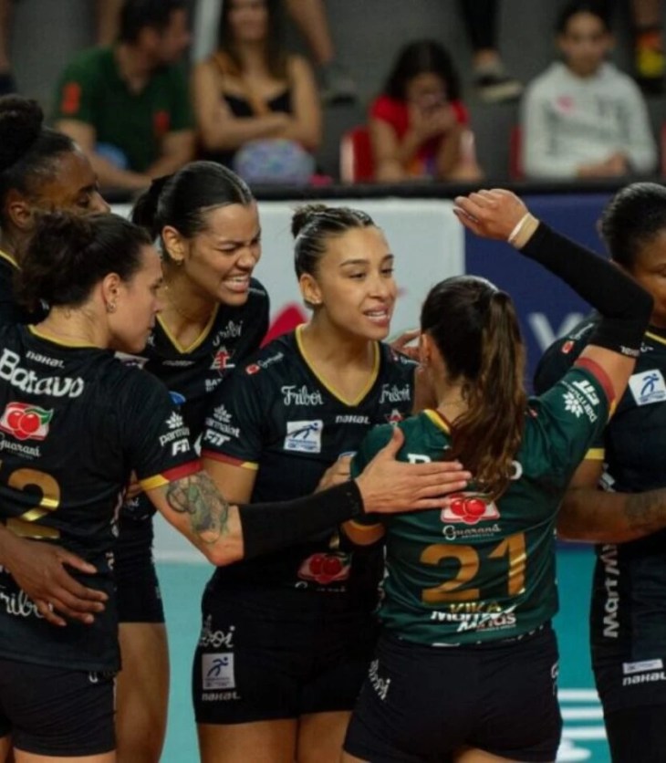 Jogadoras da equipe de vôlei feminino Batavo Mackeinze celebram juntas em quadra, abraçadas em um círculo. Vestem uniformes pretos com detalhes em amarelo e verde. A jogadora de costas, com o número 21 em verde, é abraçada pelas companheiras, enquanto outras sorriem e se apoiam mutuamente. O fundo mostra o ginásio com espectadores sentados nas arquibancadas e painéis publicitários. A cena transmite espírito de equipe, emoção e celebração após uma jogada ou ponto importante.