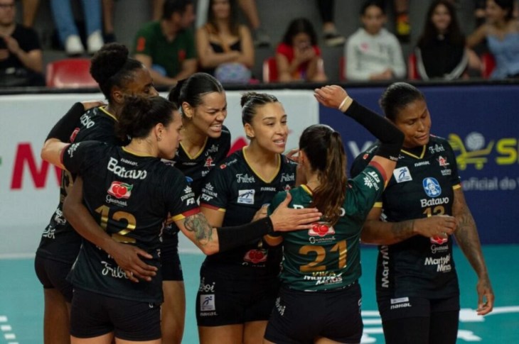Jogadoras da equipe de vôlei feminino Batavo Mackeinze celebram juntas em quadra, abraçadas em um círculo. Vestem uniformes pretos com detalhes em amarelo e verde. A jogadora de costas, com o número 21 em verde, é abraçada pelas companheiras, enquanto outras sorriem e se apoiam mutuamente. O fundo mostra o ginásio com espectadores sentados nas arquibancadas e painéis publicitários. A cena transmite espírito de equipe, emoção e celebração após uma jogada ou ponto importante.