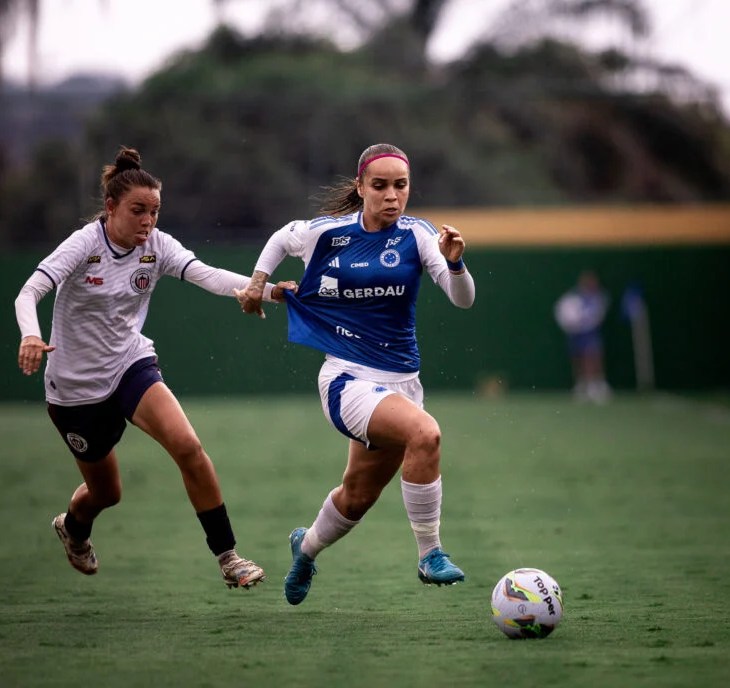 Cena de um jogo do Campeonato Mineiro Feminino entre Itabirito e o Cruzeiro. Uma jogadora do Cruzeiro, vestindo camisa azul com detalhes brancos e o patrocínio “GERDAU” no peito, corre com determinação em direção à bola branca e colorida, controlando-a com o pé direito. Ela é marcada de perto por uma atleta da Seleção de Itabirito, que usa uniforme branco com detalhes escuros e tenta desarmá-la, segurando sua camisa. Ambas estão em movimento intenso sobre o gramado verde, com fundo desfocado mostrando arquibancadas e vegetação ao longe. A imagem transmite velocidade, disputa e concentração típicas de uma partida decisiva no futebol feminino mineiro.