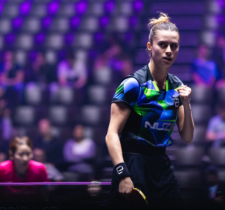 Bruna Takahashi, atleta brasileira de tênis de mesa, em ação durante uma partida. Ela está concentrada, com o punho cerrado em gesto de determinação, vestindo uniforme azul e verde com o logo 'Nuzi'. Ao fundo, arquibancadas com espectadores e iluminação roxa.