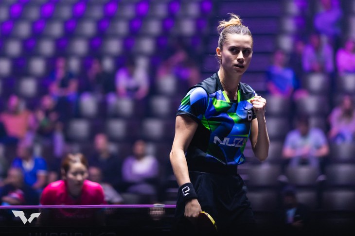 Bruna Takahashi, atleta brasileira de tênis de mesa, em ação durante uma partida. Ela está concentrada, com o punho cerrado em gesto de determinação, vestindo uniforme azul e verde com o logo 'Nuzi'. Ao fundo, arquibancadas com espectadores e iluminação roxa.