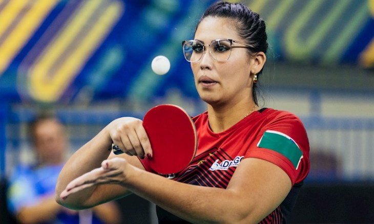 A atleta Thaís Severo em ação durante uma partida de tênis de mesa. Ela está concentrada, usando óculos de armação clara e com o cabelo preso. Veste uma camisa vermelha com detalhes pretos e listras diagonais, exibindo o patrocínio “SADOC” no peito e uma faixa verde e branca no braço esquerdo. Com a raquete vermelha na mão direita, ela prepara para sacar a bola branca. O fundo desfocado mostra o ambiente de competição, com painéis azuis e amarelos, destacando o foco da atleta no momento do jogo.