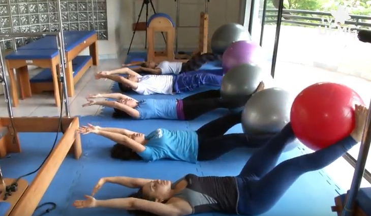 Grupo de participantes do projeto de extensão Pilates na UFMG realiza exercícios em uma sala de atividades físicas. As pessoas estão deitadas de costas sobre esteiras azuis, alinhadas em fileira, com os braços estendidos acima da cabeça e as pernas levantadas, segurando bolas suíças coloridas (vermelha, cinza e roxa) entre os pés. Ao fundo, equipamentos de Pilates, como o “reformer” de madeira e espelhos, complementam o ambiente. A sala é bem iluminada, com janelas que dão para o exterior, e o piso é claro, sugerindo um espaço universitário dedicado à saúde e ao bem-estar. A imagem transmite disciplina, concentração e o compromisso da UFMG com a promoção de qualidade de vida por meio da extensão acadêmica.