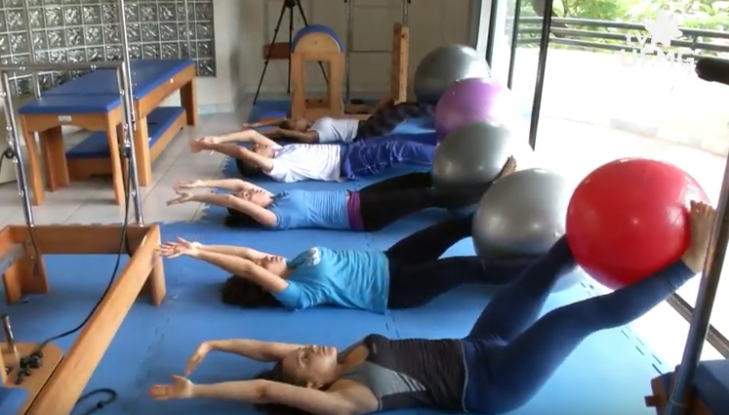 Grupo de participantes do projeto de extensão Pilates na UFMG realiza exercícios em uma sala de atividades físicas. As pessoas estão deitadas de costas sobre esteiras azuis, alinhadas em fileira, com os braços estendidos acima da cabeça e as pernas levantadas, segurando bolas suíças coloridas (vermelha, cinza e roxa) entre os pés. Ao fundo, equipamentos de Pilates, como o “reformer” de madeira e espelhos, complementam o ambiente. A sala é bem iluminada, com janelas que dão para o exterior, e o piso é claro, sugerindo um espaço universitário dedicado à saúde e ao bem-estar. A imagem transmite disciplina, concentração e o compromisso da UFMG com a promoção de qualidade de vida por meio da extensão acadêmica.
