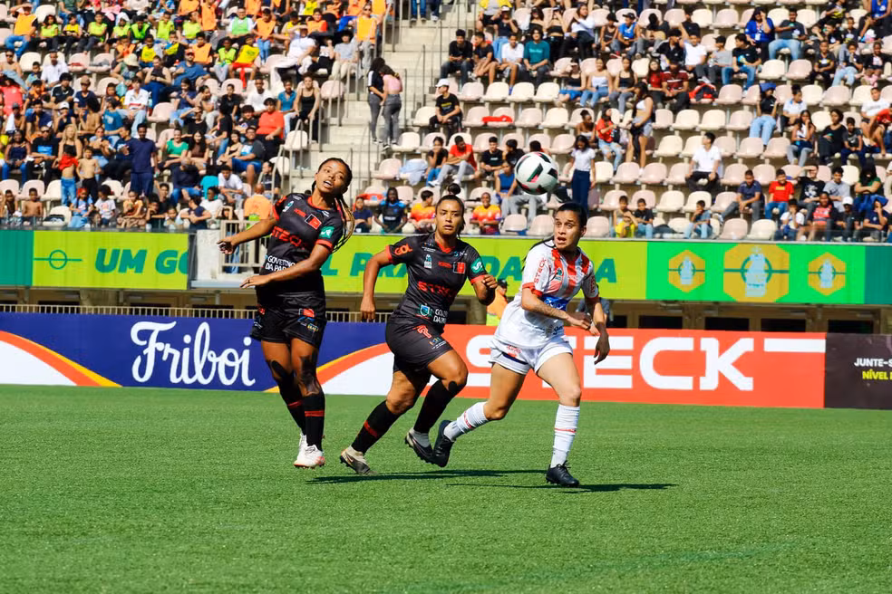 Cena de uma partida da Taça das Favelas entre a Seleção da Paraíba e Minas Gerais. Uma jogadora de Minas Gerais, vestindo uniforme branco com detalhes vermelhos, busca controlar enquanto é marcada de perto por duas adversárias da Paraíba, que usam uniformes pretos com detalhes vermelhos. O gramado verde está em destaque sob o sol, e ao fundo, arquibancadas ocupadas parcialmente por torcedores completam a atmosfera.