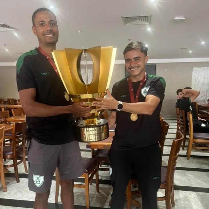 Os membros de comissão técnica Esdras e Thiago, ex-alunos do Ceca/UFMG, sorriem orgulhosos enquanto seguram juntos o troféu dourado de campeões do Campeonato Mineiro Sub-17 2025, conquistado pela equipe do América. Ambos vestem uniformes esportivos pretos com detalhes verdes - símbolo do clube - e usam medalhas douradas penduradas no pescoço. Esdras, à esquerda, veste shorts cinza e segura o troféu com ambas as mãos; Thiago, à direita, usa calça preta e apoia uma das mãos no troféu, exibindo um relógio no pulso. O fundo mostra um ambiente interno, possivelmente um hotel ou centro de convenções, com mesas, cadeiras e outros membros da comissão ao fundo. A imagem celebra a dedicação, o sucesso e a trajetória dos dois profissionais que levaram a equipe jovem ao título estadual.