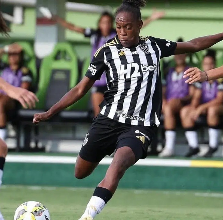 Cena de um jogo do Campeonato Mineiro Feminino entre Atlético e América. Uma jogadora do Atlético, vestindo camisa listrada preta e branca com o patrocínio “H2bet” no peito, chuta a bola com o pé direito. O gramado verde está bem cuidado, e ao fundo, o banco de reserva de uma das equipes com atletas sentadas em cadeiras que completam o cenário. A imagem captura a concentração e habilidade de um clássico mineiro no futebol feminino.