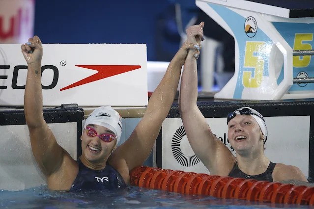 A nadadora Mafê Costa, com touca branca e óculos de natação rosa, celebra na piscina após quebra de record nacional nos 200m livre. Ela ergue os dois braços em gesto de vitória, sorrindo, enquanto está parcialmente imersa na água. Ao seu lado, outra atleta também comemora, segurando a mão de Mafê em um momento de alegria. Ao fundo, vê-se o painel da raia 5, o logo da marca “Speedo” e parte da estrutura de salto. A cena transmite emoção, conquista e espírito esportivo na competição de natação.