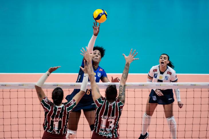 Cena dinâmica de uma partida de vôlei feminino entre o Gerdau Minas e o Fluminense. Uma jogadora do Minas, vestindo camisa azul, salta acima da rede para atacar a bola amarela e azul. Do lado oposto, duas jogadoras do Fluminense - Lara, com o número 1, e Ariane, com o número 13 -, ambas em uniformes listrados em verde e grená, saltam juntas em tentativa de bloqueio, com os braços estendidos. Ao fundo, outra atleta do Minas, em camisa branca, observa a jogada. A rede exibe o logo “VÔLEI BRASIL”. O fundo é composto pelo piso da quadra azul e rosa claro.