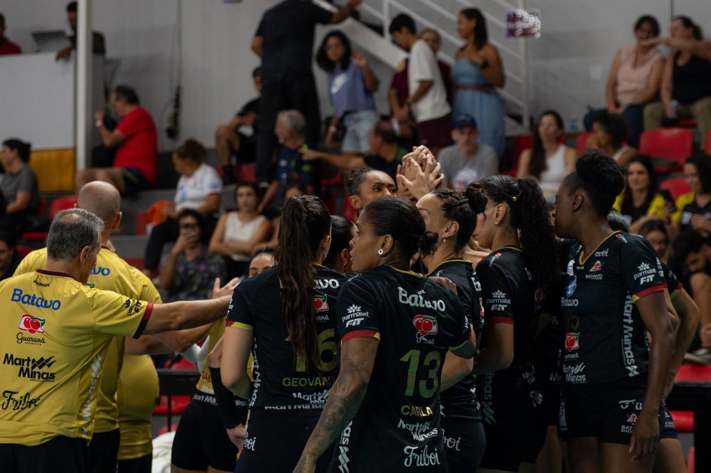 A women's volleyball team huddled together in a gymnasium, celebrating and strategizing during a match, with spectators visible in the background.
